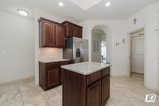 kitchen with arched walkways, light stone counters, a kitchen island, decorative backsplash, and stainless steel fridge