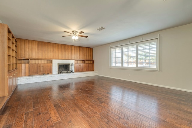 unfurnished living room featuring wood walls, ceiling fan, dark wood-type flooring, and a brick fireplace