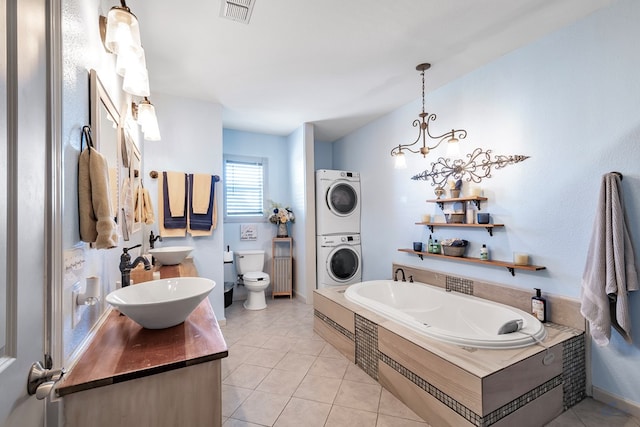 bathroom with an inviting chandelier, tile patterned flooring, a tub to relax in, toilet, and stacked washer and clothes dryer