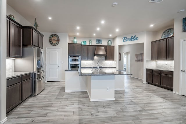 807 Bronco Ct, Midland, TX 79705 - kitchen with custom exhaust hood, tasteful backsplash, dark stone counters, dark brown cabinets, and an island with sink