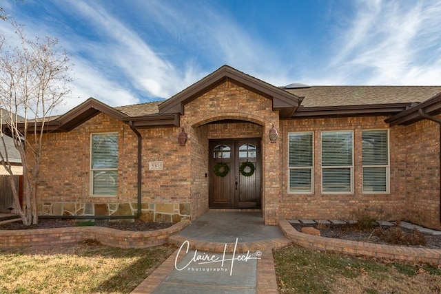 view of exterior entry featuring french doors, roof with shingles, and brick siding