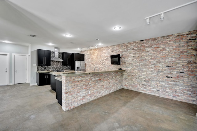 kitchen with concrete flooring, stainless steel refrigerator with ice dispenser, wall chimney range hood, and dark cabinetry
