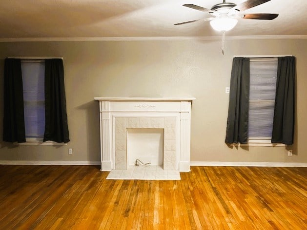 unfurnished living room featuring ceiling fan, a fireplace, baseboards, hardwood / wood-style floors, and crown molding