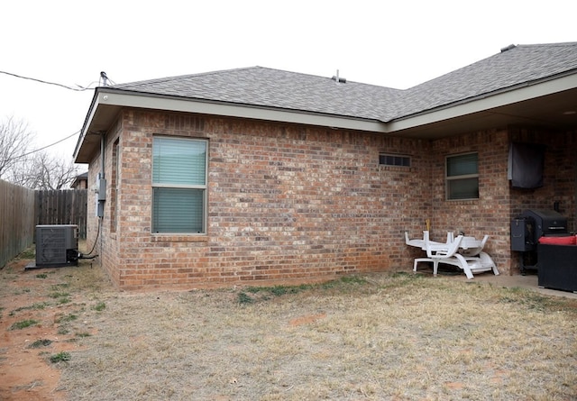 view of property exterior with central air condition unit, roof with shingles, fence, and brick siding