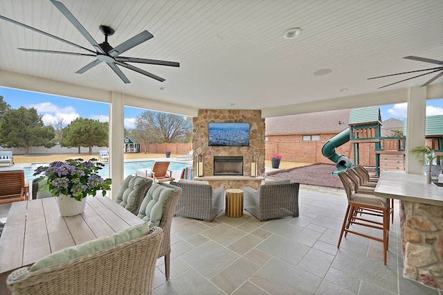 view of patio featuring fence, an outdoor stone fireplace, outdoor dry bar, an outdoor pool, and ceiling fan