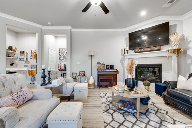 living room featuring crown molding, a ceiling fan, light wood finished floors, a brick fireplace, and visible vents