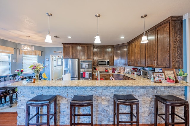 kitchen with stainless steel appliances, light stone countertops, a peninsula, hanging light fixtures, and dark brown cabinets