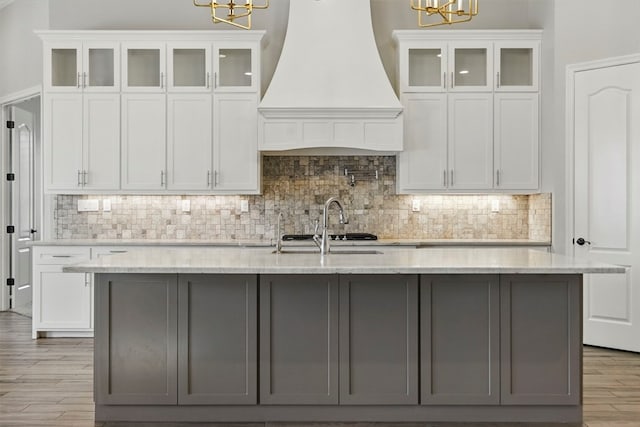 kitchen featuring custom range hood, a chandelier, a center island with sink, and white cabinetry