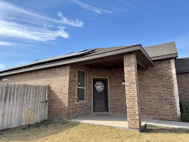 view of exterior entry with a lawn, a patio, fence, roof mounted solar panels, and brick siding