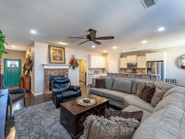 living room featuring ceiling fan, a brick fireplace, dark hardwood / wood-style floors, and a textured ceiling