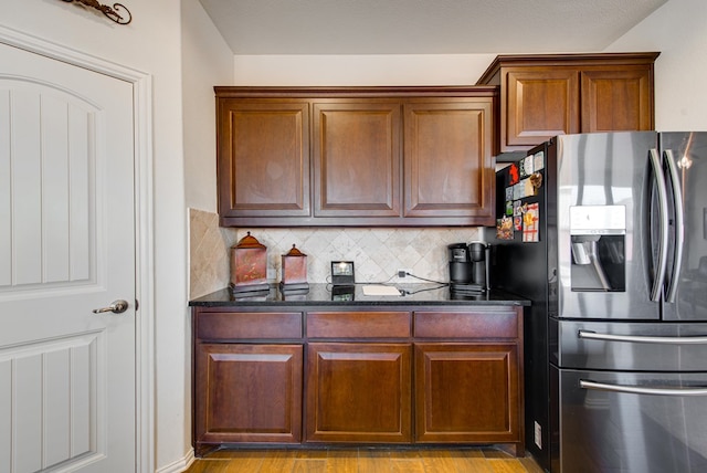 kitchen with brown cabinetry, dark stone counters, light wood-type flooring, stainless steel refrigerator with ice dispenser, and backsplash