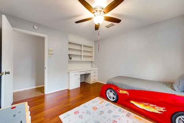 bedroom with wood finished floors, ceiling fan, built in study area, baseboards, and visible vents
