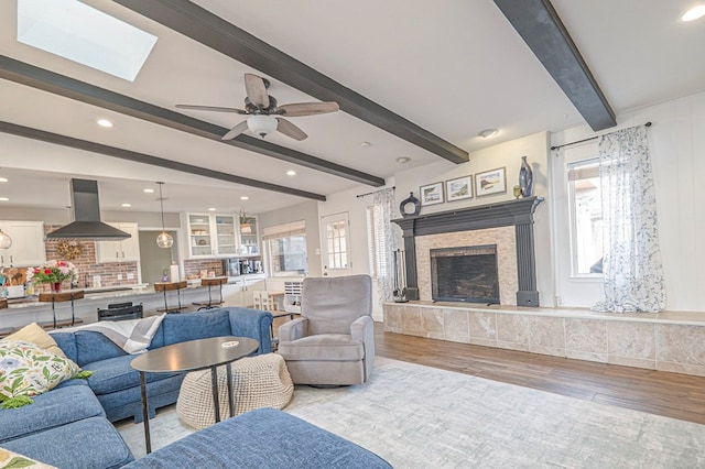 living room with a skylight, a tiled fireplace, light wood-style flooring, ceiling fan, and beam ceiling