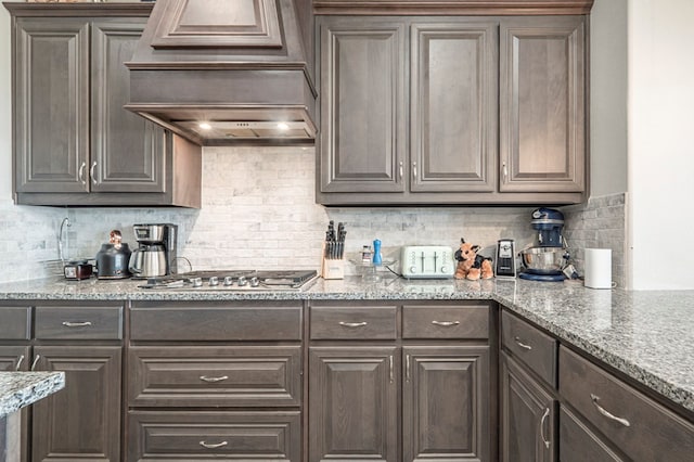 kitchen with stainless steel gas cooktop, dark brown cabinets, premium range hood, light stone counters, and backsplash
