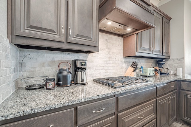 kitchen featuring decorative backsplash, stainless steel gas cooktop, dark brown cabinets, premium range hood, and light stone counters