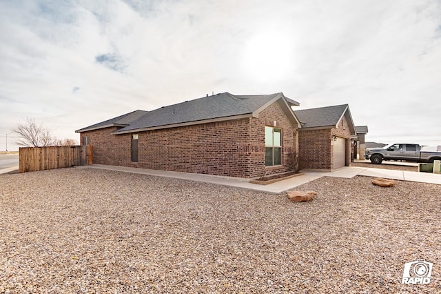 view of side of property featuring a garage, brick siding, fence, driveway, and roof with shingles