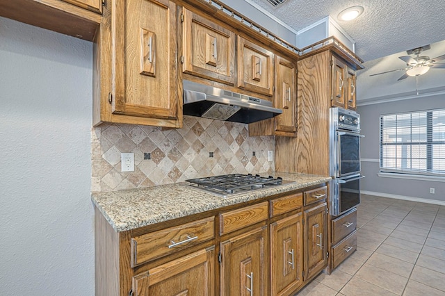 kitchen featuring brown cabinets, multiple ovens, under cabinet range hood, stainless steel gas cooktop, and light tile patterned flooring