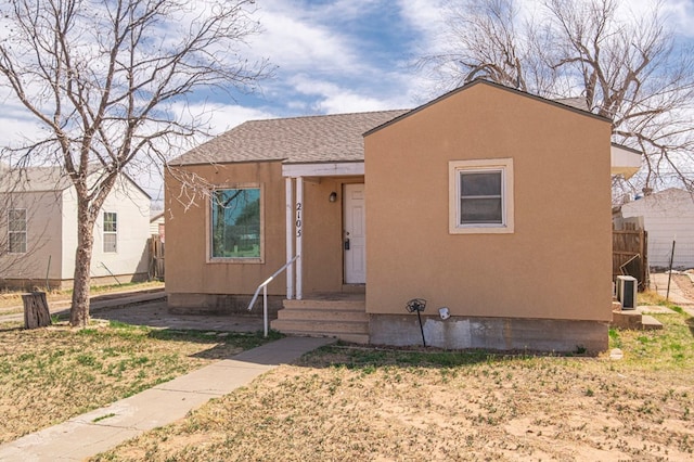 bungalow-style home with a shingled roof, stucco siding, fence, and central air condition unit