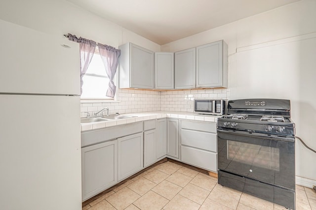 kitchen featuring tile counters, stainless steel microwave, black gas range, freestanding refrigerator, and a sink