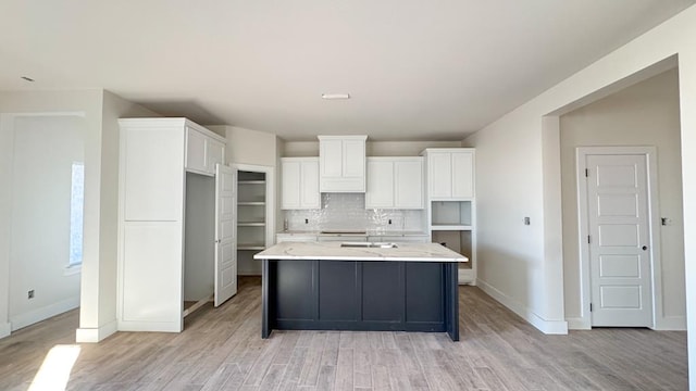 kitchen with backsplash, light stone countertops, an island with sink, white cabinets, and light wood-type flooring