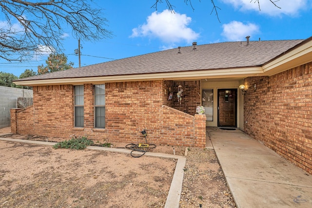 view of exterior entry with roof with shingles, fence, and brick siding