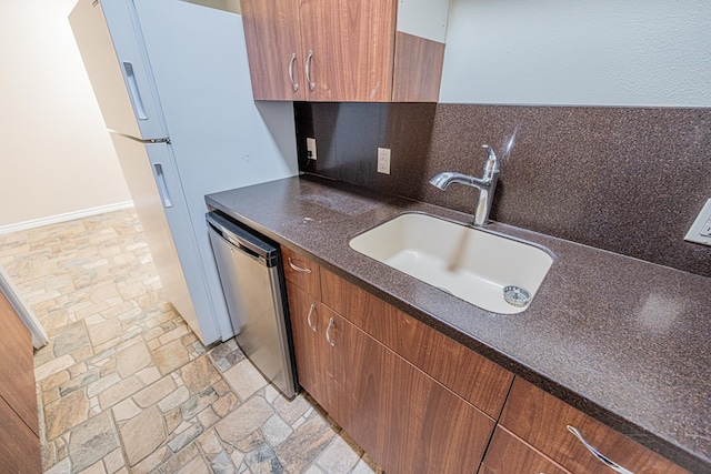 kitchen with a sink, stainless steel dishwasher, freestanding refrigerator, brown cabinets, and stone tile flooring