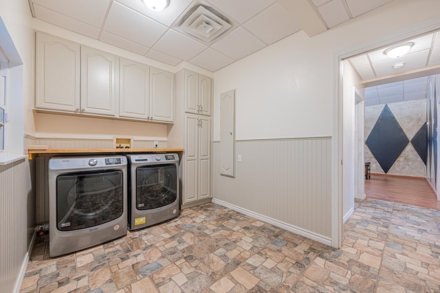 laundry area featuring a wainscoted wall, baseboards, cabinet space, stone finish floor, and washing machine and clothes dryer