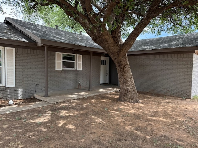 property entrance featuring brick siding, a shingled roof, and a patio