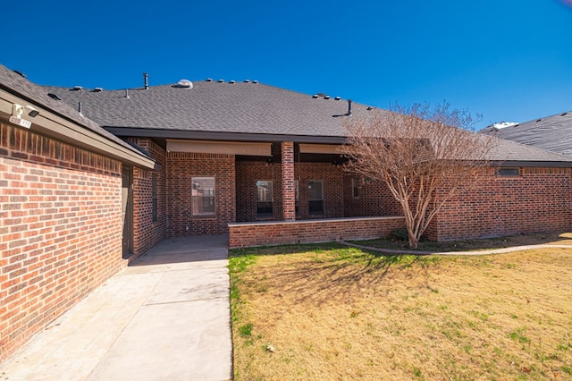 entrance to property featuring brick siding, roof with shingles, a patio, and a yard