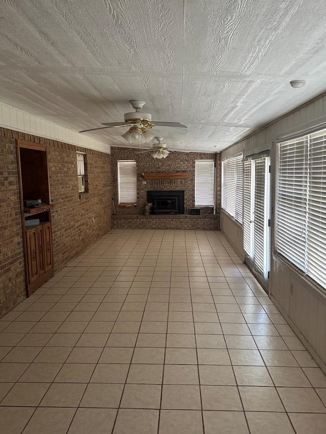 unfurnished living room featuring a brick fireplace, light tile patterned flooring, ceiling fan, a textured ceiling, and brick wall