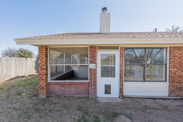 view of exterior entry with brick siding, a chimney, a shingled roof, and fence