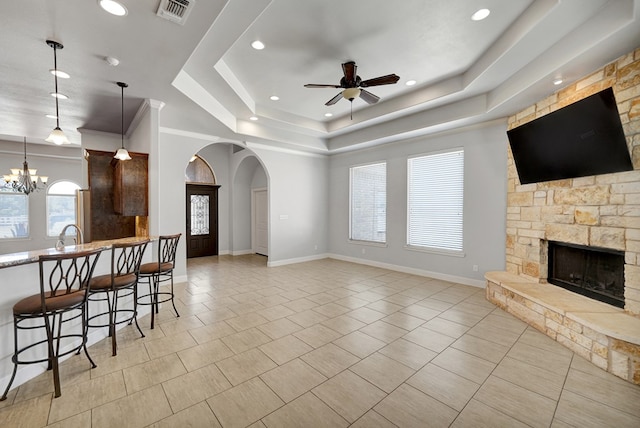 living room with ceiling fan with notable chandelier, arched walkways, a stone fireplace, visible vents, and a raised ceiling