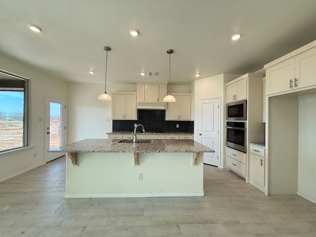 kitchen featuring tasteful backsplash, a center island with sink, appliances with stainless steel finishes, light stone countertops, and a sink