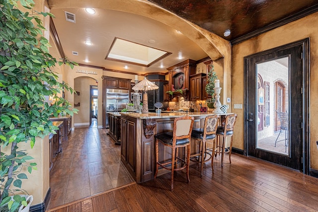 kitchen with arched walkways, a breakfast bar, dark wood-style floors, ornamental molding, and light stone counters