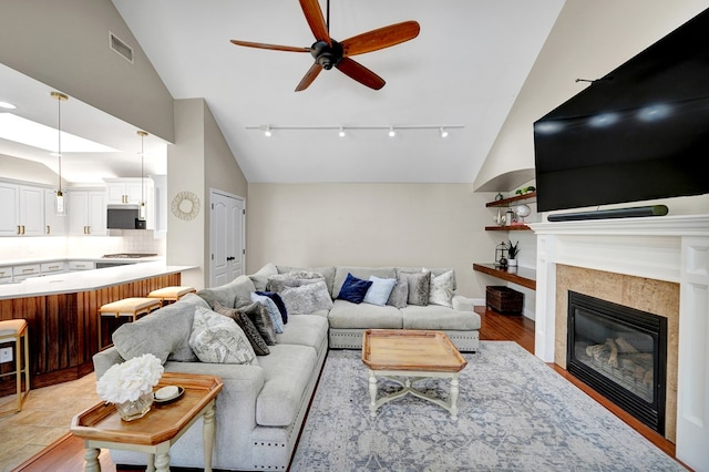 living room with ceiling fan, rail lighting, a tiled fireplace, high vaulted ceiling, and light wood-type flooring