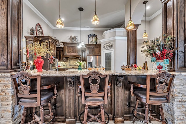 kitchen with dark brown cabinetry, stainless steel fridge, light stone counters, and ornamental molding
