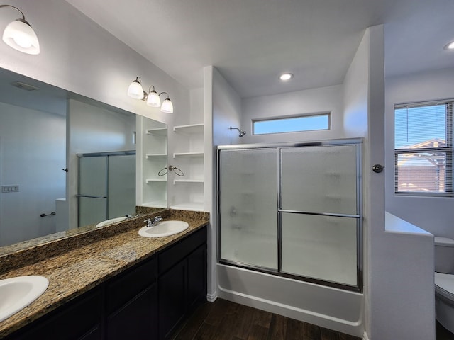 full bathroom with toilet, a sink, wood finished floors, double vanity, and bath / shower combo with glass door