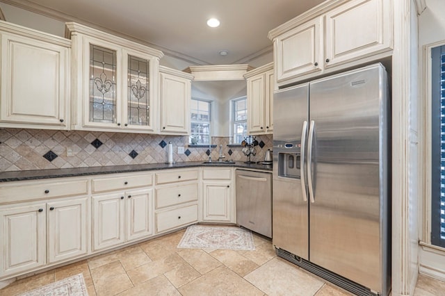 kitchen with glass insert cabinets, a sink, cream cabinets, stainless steel appliances, and backsplash