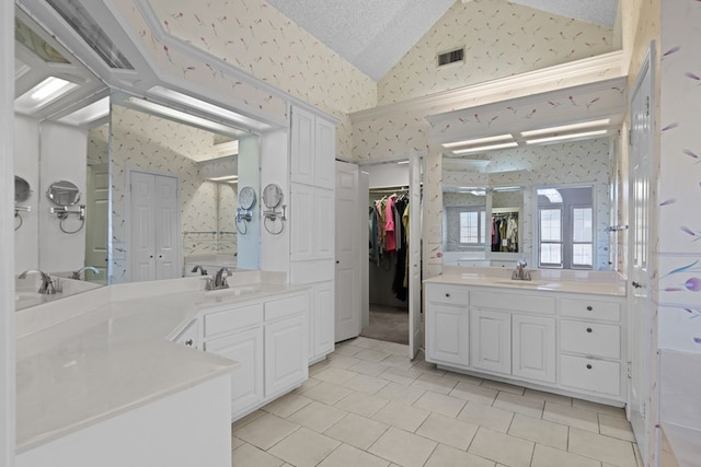 bathroom featuring visible vents, two vanities, a sink, wallpapered walls, and vaulted ceiling