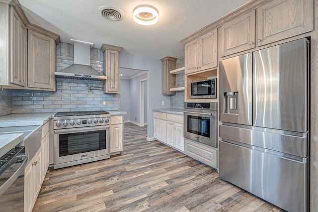 kitchen with wall chimney range hood, appliances with stainless steel finishes, wood-type flooring, decorative backsplash, and light brown cabinets