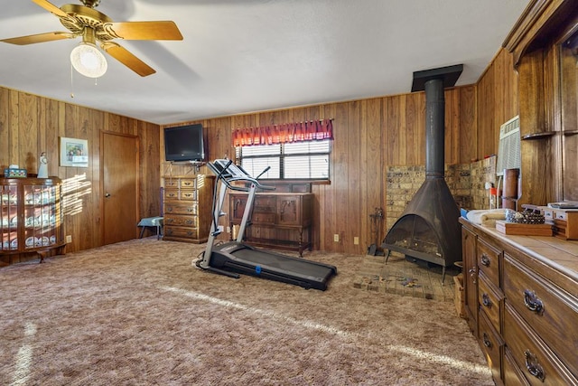 exercise room with carpet, a wood stove, ceiling fan, and wooden walls