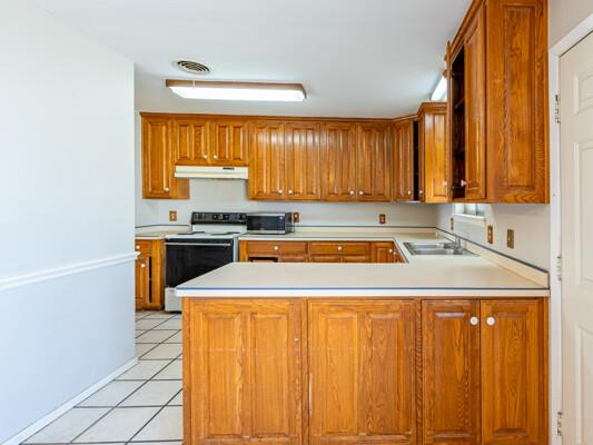 kitchen featuring white electric stove, under cabinet range hood, a peninsula, light countertops, and brown cabinetry