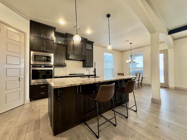 kitchen with dark brown cabinets, stainless steel appliances, decorative light fixtures, light hardwood / wood-style flooring, and a center island with sink