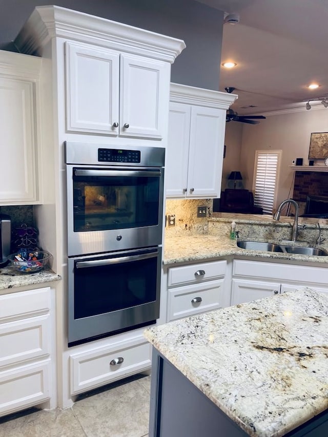 kitchen with double oven, a sink, white cabinetry, backsplash, and light stone countertops