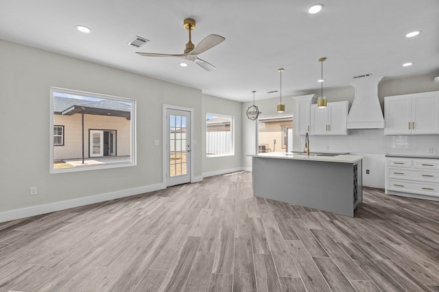 kitchen featuring tasteful backsplash, custom exhaust hood, light hardwood / wood-style flooring, white cabinets, and an island with sink