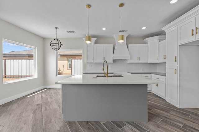 kitchen with custom exhaust hood, a kitchen island with sink, a healthy amount of sunlight, white cabinets, and hanging light fixtures