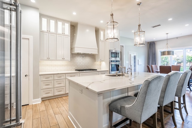 15 Lakes Dr, Midland, TX 79705 - kitchen with custom range hood, appliances with stainless steel finishes, light wood finished floors, tasteful backsplash, and a center island with sink