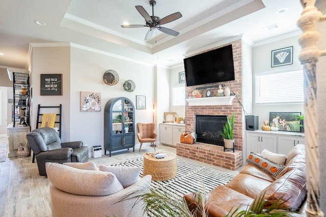 living room featuring ceiling fan, a brick fireplace, a raised ceiling, ornamental molding, and light wood-type flooring