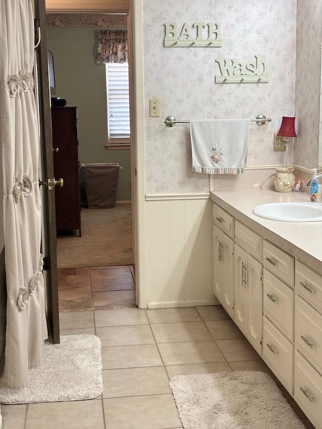 bathroom featuring tile patterned flooring, vanity, wainscoting, and wallpapered walls