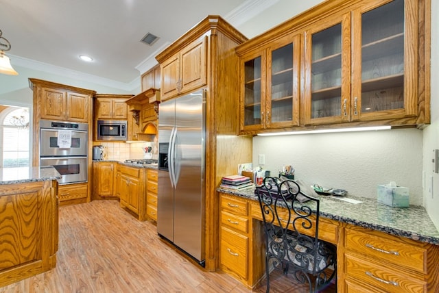 kitchen featuring dark stone counters, brown cabinets, built in appliances, crown molding, and built in desk